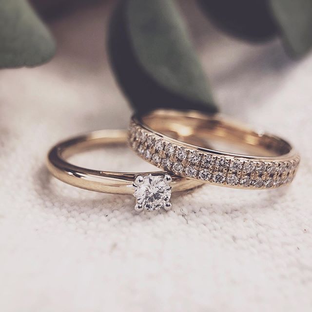 Twee gouden ringen rusten op een lichte stof: de Excellent Jewelry Bicolor Wit- en Geelgouden Solitair met Briljant 0.19 crt. en een trouwring met diamant. Zachtgroene bladeren zijn vaag op de achtergrond.