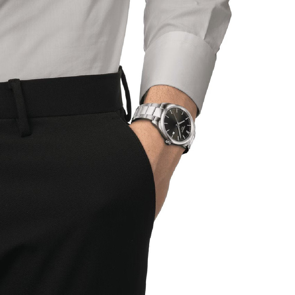 Een persoon met zijn hand in zijn zak draagt een zwarte broek, een lichtgrijs shirt met lange mouwen en een PR 100-horloge van Tissot met een zwarte wijzerplaat.