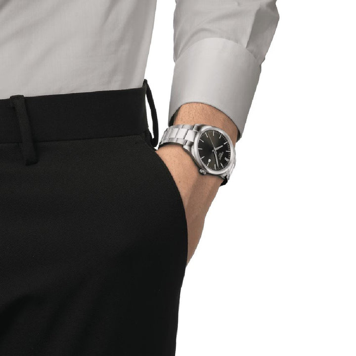 Een persoon met zijn hand in zijn zak draagt een zwarte broek, een lichtgrijs shirt met lange mouwen en een PR 100-horloge van Tissot met een zwarte wijzerplaat.